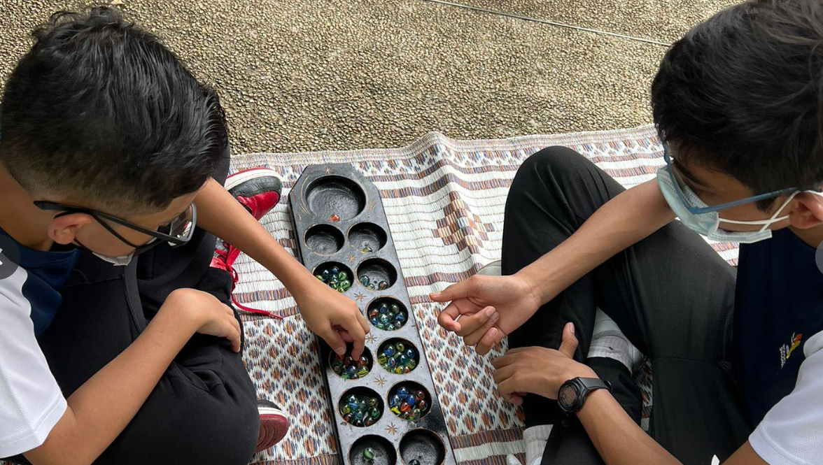 Two masked boys sitting on a mat playing congkak with colorful marbles in a wooden board.